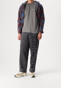 Flanellen overhemd met rood, blauw en zwart geruit patroon over een grijs T-shirt met 'obey' logo. Donkergrijze wijde broek met zwarte grafische print, beige sneakers.