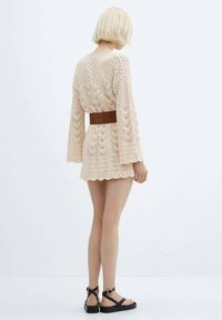 Crème-kleurige gehaakte jurk met klokmouwen, golvende zoom en een bruine riem in de taille. Gemaakt met een paar zwarte sandalen met bandjes.