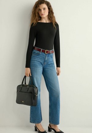 Młoda kobieta w czarnej bluzce z długim rękawem, niebieskich jeansach z brązowym paskiem, czarnych butach na obcasie, trzymająca czarną skórzaną torebkę, stoi na tle białej ściany.