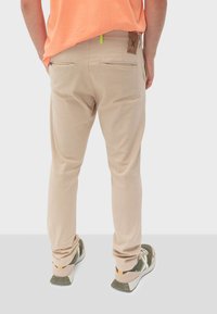 Pantaloni beige con una vestibilità slim, caratterizzati da due tasche posteriori con zip e un'etichetta in pelle marrone. Accoppiati con sneaker multicolori.