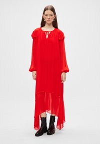 Vestido maxi rojo y transparente con mangas acampanadas y cuello con lazo. Presenta un dobladillo asimétrico y se combina con botas negras hasta los tobillos.