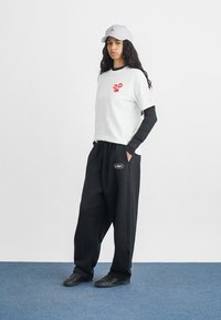 Λευκό T-shirt με γραφικό κόκκινων χειλιών, τοποθετημένο πάνω από μαύρο μπλουζάκι μέχρι τα μανίκια; χαλαρές μαύρες φόρμες με λογότυπο; γκρι καπέλο και μαύρα παπούτσια.