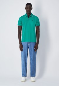 Grünes Poloshirt mit schwarzem Kragen und weißen Paspeln, kombiniert mit hellblauen Jeans und weißen Sneakern. Slim-Fit und mit kurzenÄrmeln.
