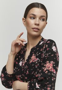 Blusa nera floreale con motivi di fiori rosa e bianchi, scollatura a V e maniche a sbuffo con polsini elasticizzati. Tessuto morbido.