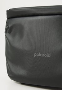 Černá voděodolná taška s hladkým povrchem a rolovacím designem, na přední straně s jemným logem Polaroid.