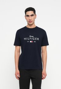 Mladý muž oblečený v námořnickém modrém tričku Tommy Hilfiger s logem a textem "1985", stojící před jasně bílým pozadím.
