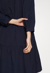 Marinblå klänning med en rynkad kjol, långa ärmar med knäppte manschetter och lätt, slätt tyg. Händerna är placerade nära midjan.