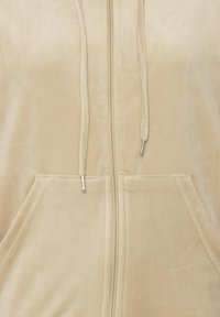 Sweat à capuche beige zippé avec poches frontales et cordons munis d'embouts métalliques sur un tissu doux.