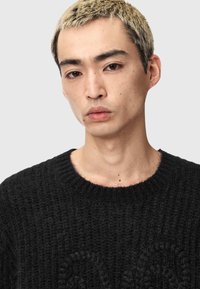 Zwarte gebreide trui met een ronde halslijn, een gestructureerd patroon en een losse pasvorm. Model heeft kort blond haar en een neutrale achtergrond.