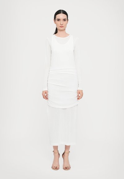 White long-sleeve dress with layered design; features a round neckline and straight cut. Model wears neutral-tone sandals. Simple texture.