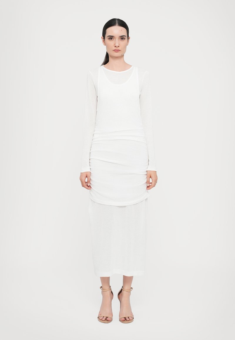 Witte lange mouw jurk met gelaagd ontwerp; heeft een ronde halslijn en rechte snit. Model draagt sandalen in neutrale tinten. Eenvoudige textuur.