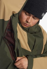 Chaqueta verde y marrón con apertura de medio cierre, forro de malla y tejido texturizado; combinada con un gorro negro; con costuras detalladas.