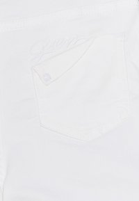 Valkoinen farkkukangas, jossa on läppätasku ja neppari. "Guess" on kirjailtu valkoisena lähelle taskua, sulautuen materiaaliin.