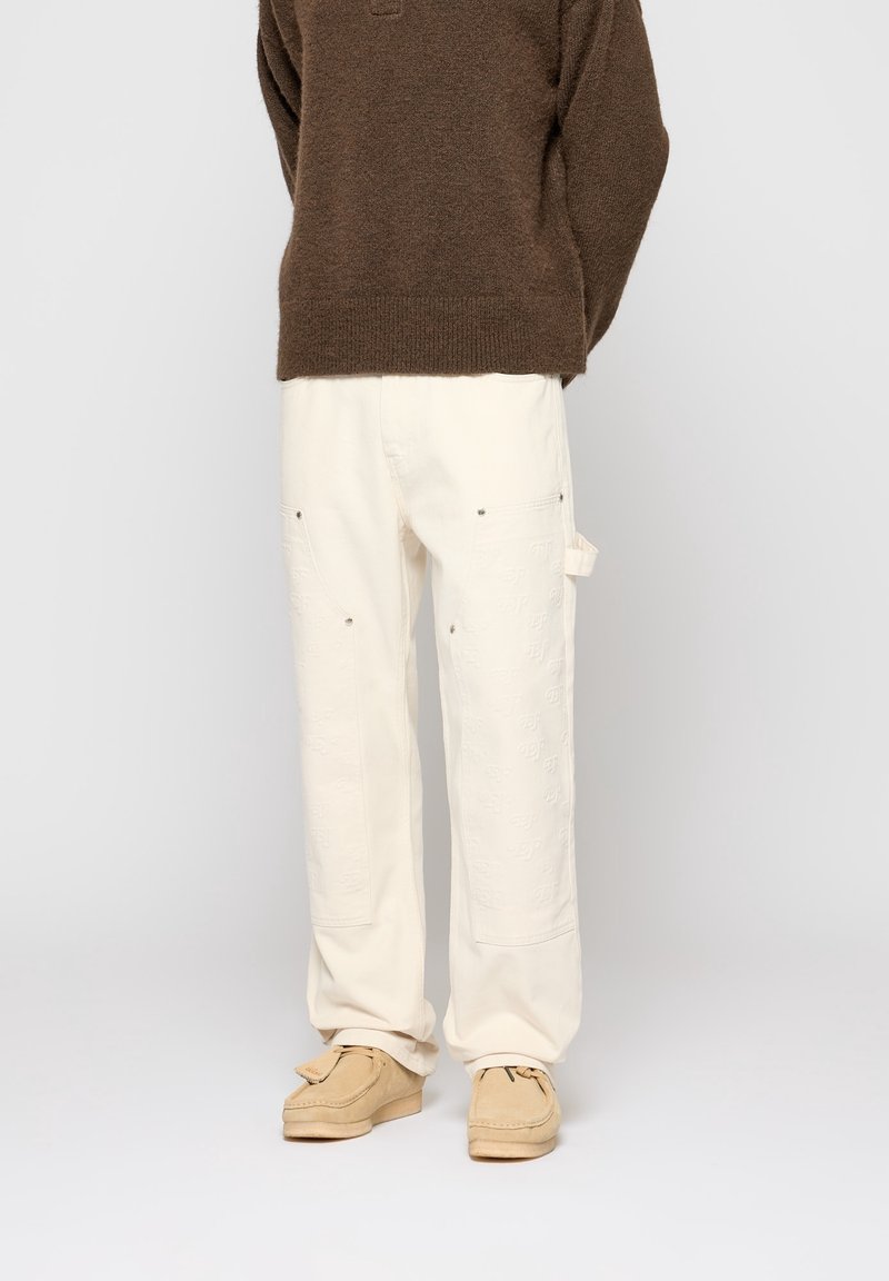 Personne portant un pull marron, un pantalon blanc ample avec des détails cousus, et des chaussures de style mocassin beige, debout devant un fond uni.