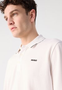 Polo triko v světle krémové barvě s texturovanou tkaninou, s černým logem "HUGO" na levé straně hrudi a kontrastním lemem límce.