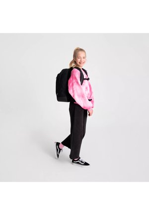 Ragazza sorridente con coda di cavallo bionda che indossa una felpa rosa tie-dye, pantaloni neri, scarpe da ginnastica e zaino nero mentre cammina su sfondo bianco.