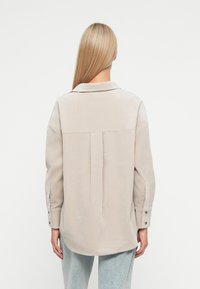 Jasnoszara koszula zapinana na guziki z plisą na plecach, z długimi rękawami i klasycznym kołnierzykiem. Miękka tekstura, luźny krój i mankiety na jeden guzik.