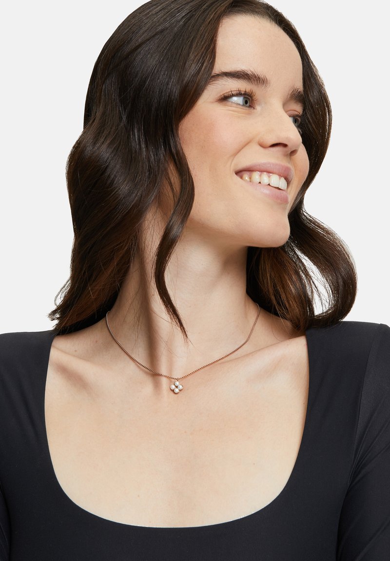 Roze gouden ketting met een bloem pendant met vier witte edelstenen. Strakke ketting, eenvoudig ontwerp, gecombineerd met een zwarte top en natuurlijk haar.