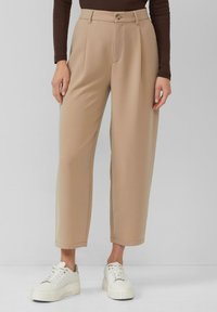 Beige maßgeschneiderte Hose mit weitem Bein und Bügelfalte, ausgestattet mit einem vorderen Knopfverschluss. Kombiniert mit weißen Turnschuhen für einen lässigen Look.