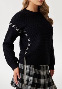 Zwarte gebreide trui met een textuurpatroon, zilveren decoratieve vetersluiting op de mouwen en een aangesloten zoom. Gecombineerd met een geruite rok.