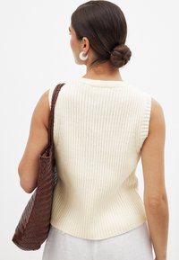 Rippenloses, ärmelloses Stricktop in Off-White mit abgerundetem Saum. Begleitet von einer braunen, gewebten Tragetasche mit breitem Trageriemen.
