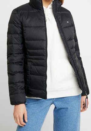 Kobieta ubrana w czarną pikowaną kurtkę na białej bluzie z kapturem i jasnoniebieskich dżinsach, prezentująca przedni zamek błyskawiczny z trójkolorową zawieszką.