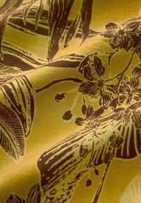 Tissu en coton avec un fond jaune orné d'un motif floral et de feuilles brun foncé, mettant en valeur des détails et textures complexes.