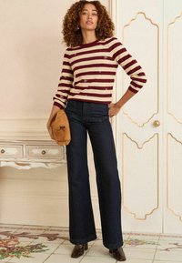 Pulover tricotat stripat în nuanțe de burgundy și crem, cu mâneci scurte, asortat cu pantaloni evazați din denim închis. O poșetă bej completează ținuta.