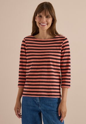 Femme souriante portant un t-shirt à manches longues rayé horizontalement corail et noir et un jean bleu denim sur un fond neutre.