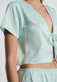 Top corto de color verde menta con escote en V, mangas cortas y tejido con estampado de corazones. Presenta un detalle de lazo en el frente y una textura suave.