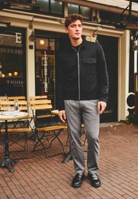 Black zip-up jacket with two chest pockets, gray textured trousers, and black loafers. Set against a café backdrop with wooden chairs.