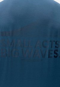 Niebieska koszulka z krótkim rękawem, wykonana z miękkiego materiału; posiada wypukły czarny napis: "MAŁE CZYNNOŚCI WIELKIE FALI" oraz subtelną grafikę fali powyżej napisu.