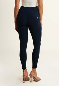 Navy Leggings mit hohem Bund, glatter Textur und ausgeprägten Nahtdetails für eine verbesserte Form. Kombiniert mit transparenten, hohen Sandalen.