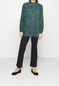 Långärmad turkos blus med ett svart leopardmönster, matchad med svarta figurnära byxor och platta skor. Slät tyg, avslappnad passform.
