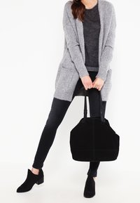 Femme portant un cardigan gris, un pull sombre, un jean noir et des bottines noires, tenant un grand sac à main noir par ses poignées.