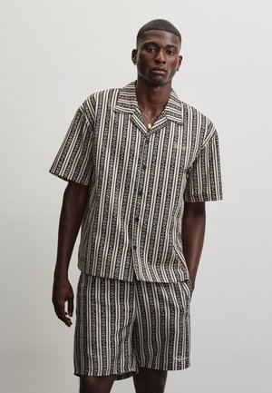 Homme portant une chemise à manches courtes boutonnée à rayures verticales noires et beiges avec un short assorti à rayures, sur un fond uni.