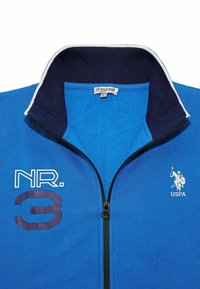 Blå zip-up sweatshirt med svart krage, prydd med vit brodyr och en grafisk siffra 3 i mörkblått. Tillverkad av mjukt tyg.