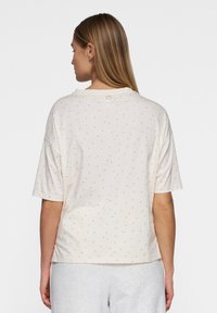 Lyscremet t-shirt med korte ærmer, prydet med et diskret mønster af guldprikket og en rund halsudskæring. Bagfra ses et lille mærke ved halsen.