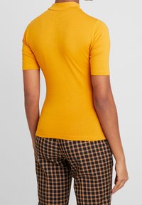 Polo tričko s krátkým rukávem v jasně žluté barvě, vyrobené z hladké tkaniny s přiléhavým střihem. Nosené s kostkovanými kalhotami v tmavých tónech.