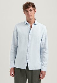Camisa de manga larga de color azul claro, con cuello abotonado, hecha de lino, que presenta una tela ligeramente texturizada y un dobladillo recto.