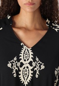 Prenda negra de cuello en V que presenta intrincados patrones florales y de cachemira en blanco, con una textura suave y lisa y detalles en los hombros.