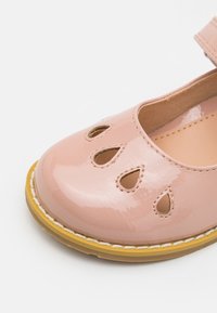 Zapato de charol rosa con punta redonda, recortes decorativos, detalles cosidos y suela de goma amarilla. Correa ajustable en el lado.