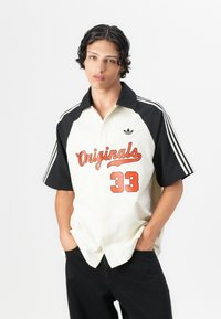 Short-sleeve button-up shirt in cream and black, featuring red text "Originals" and number "33." Black shoulder stripes and logo detail.