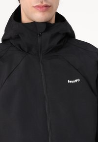 Black zip-up jacket with a hood. Features a smooth texture and a white logo on the chest, with subtle seam detailing on shoulders.