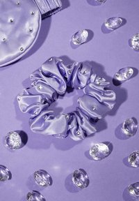 Fioletowy satynowy scrunchie z srebrnymi ćwiekami, otoczony przez przezroczyste dekoracyjne kamienie na pasującym fioletowym tle.