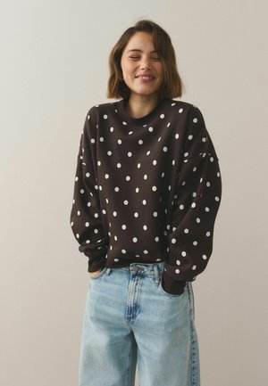 Rövid barna hajú nő, sötétbarna pöttyös pulóvert és világoskék farmert visel, mosolyogva, becsukott szemmel és zsebre tett kézzel.