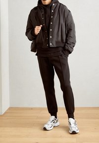 Svart kort bomberjacka med silverfärgade tryckknappar, lagd ovanpå en svart hoodie. Svarta avsmalnande byxor och silverfärgade sportskor med detaljer.