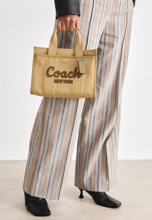 Tan värvi Coachi kott, millel on tikitud "Coach New York" logo, kuldne riistvara, kaks sangat ja silt. Kombineeritud triibuliste laia säärega pükstega.