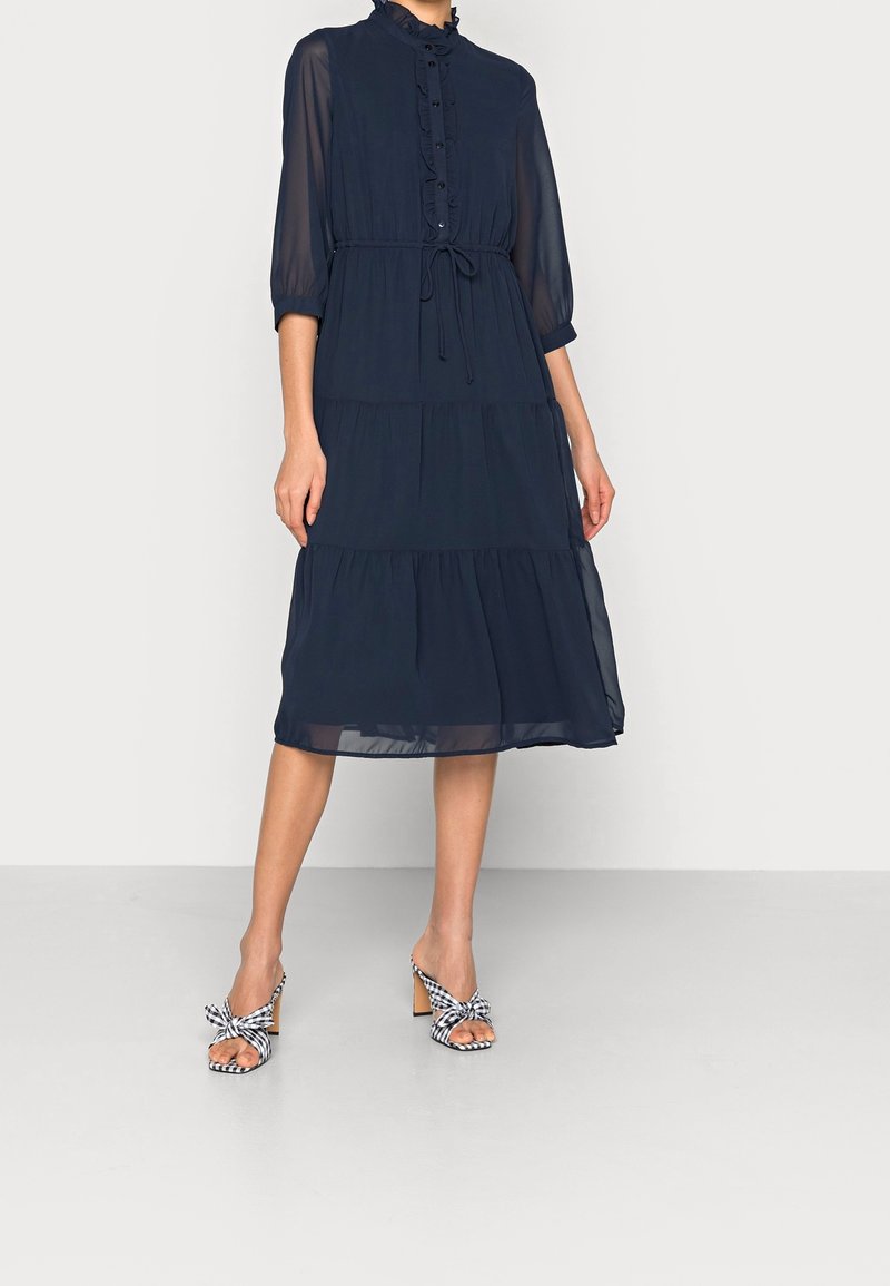 Námořnickomodré šaty s polovičními rukávy, volánkovými knoflíky, stahováním v pase a vrstveným designem, doplněné černobílými kostkovanými sandály na podpatku.