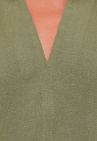 Olivengrøn hørbluse med dyb V-udskæring og diskrete syningsdetaljer, som fremviser et tekstureret stof i et nærbillede.